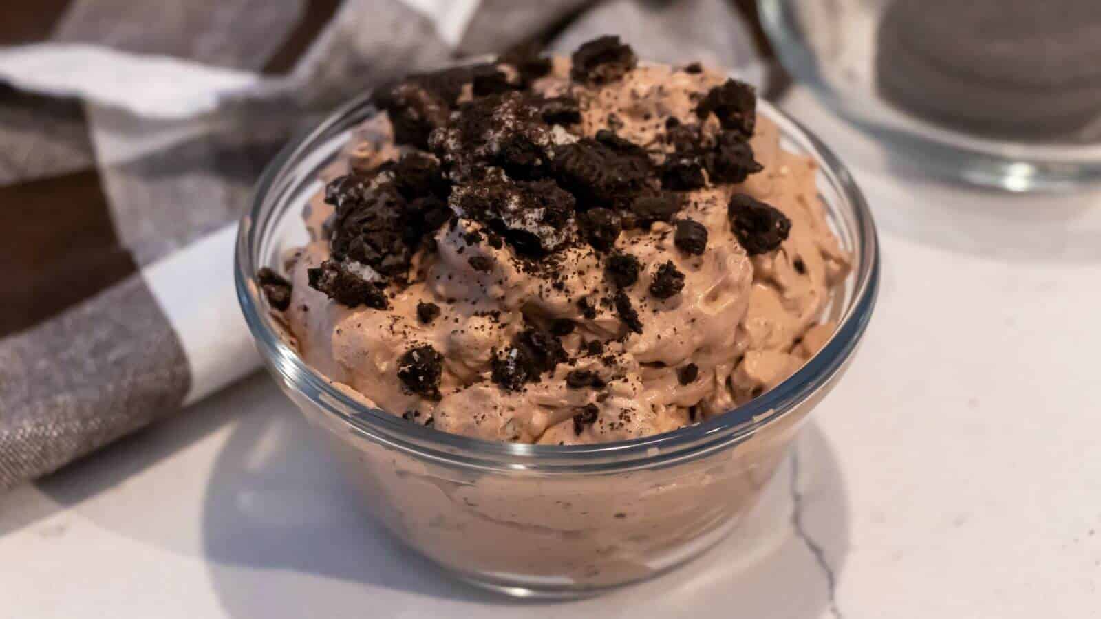 A glass bowl filled with chocolate whipped dessert topped with crushed chocolate cookies sits on a white surface near a brown and white cloth.
