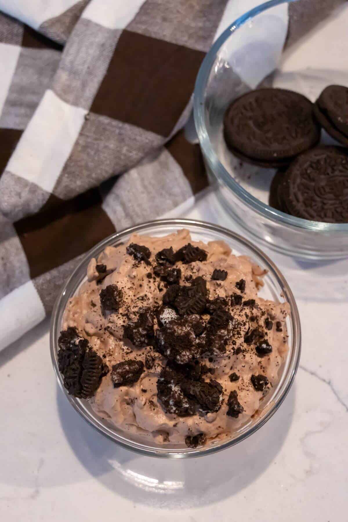 A glass bowl filled with cookies and cream-flavored dessert topped with crushed chocolate sandwich cookies, next to a bowl of whole cookies and a checkered cloth.