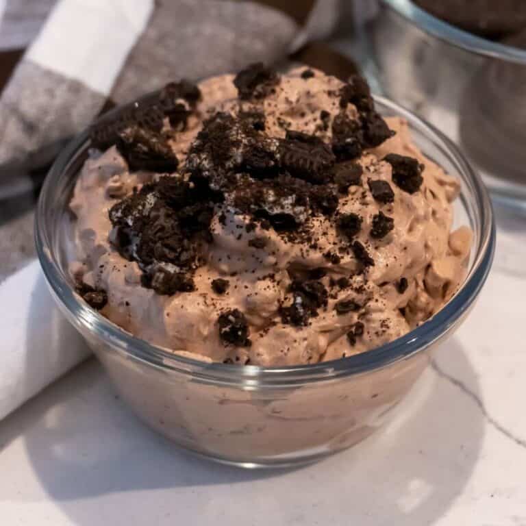 A glass bowl filled with chocolate mousse topped with crushed chocolate cookies, placed on a white surface near a cloth napkin.