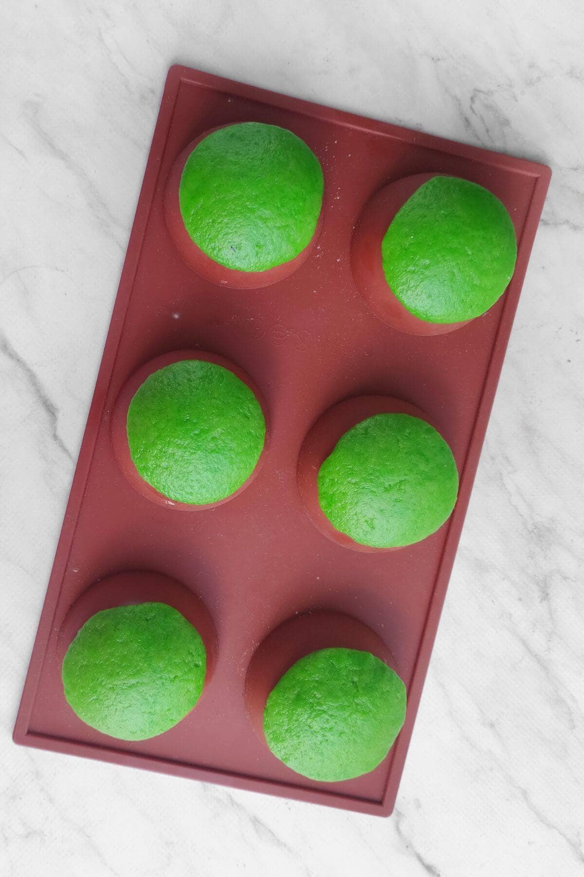 Six green-topped round buns in a red silicone baking mold are placed on a white marble surface.