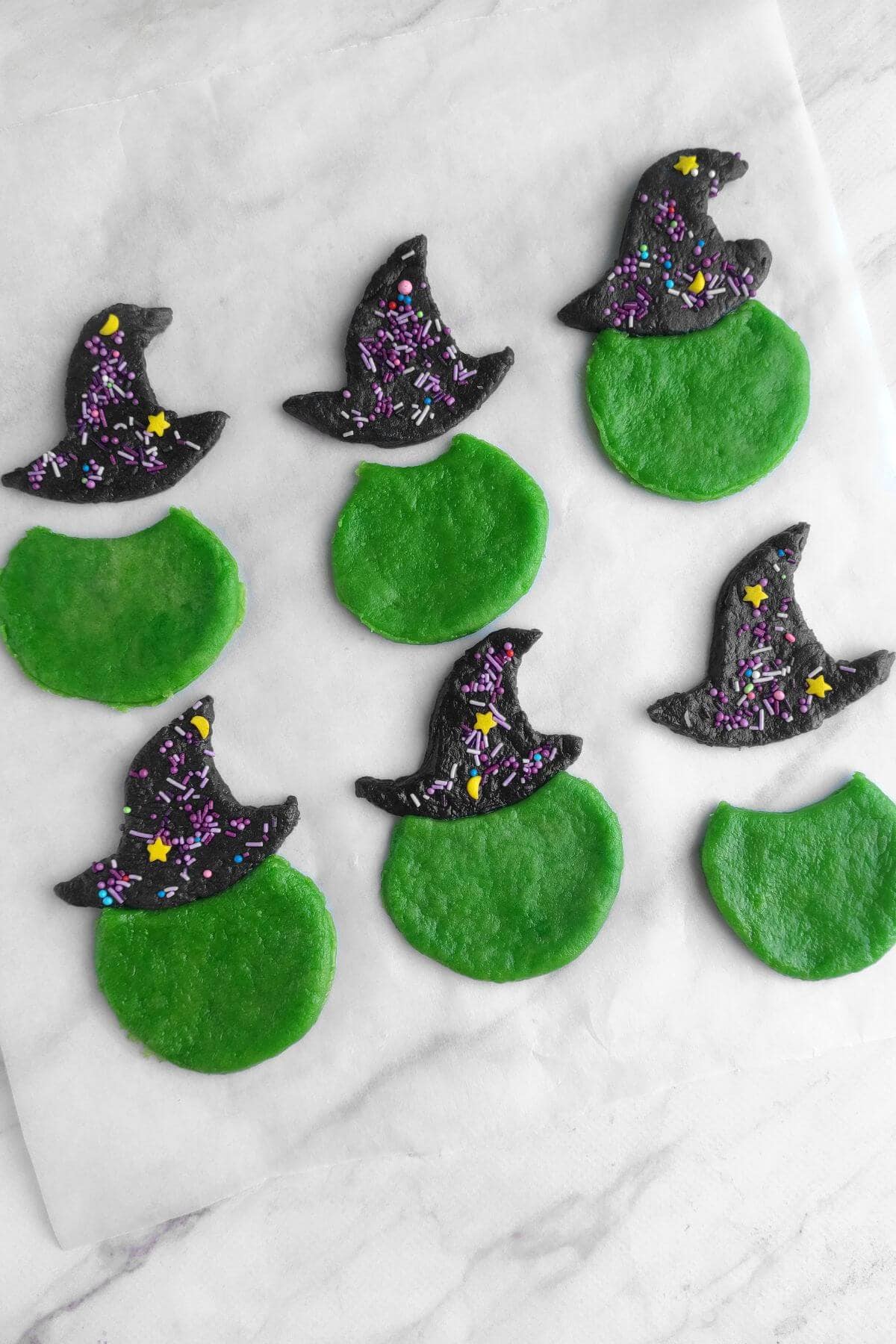 Six unbaked cookies on parchment, each consisting of a round green base and a black hat-shaped top decorated with purple sprinkles and star-shaped confetti.