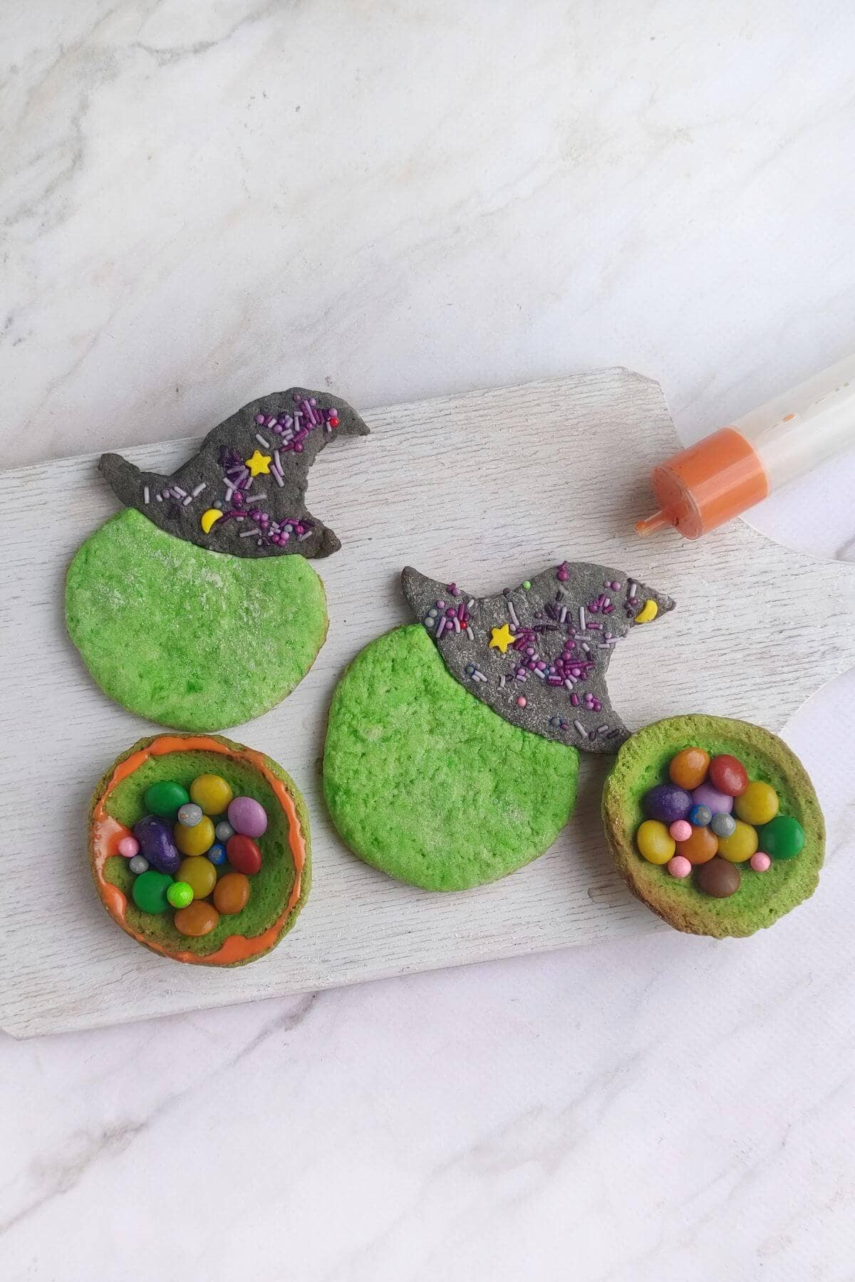 Two green cookies shaped like witch hats and two green cookies filled with colorful candies are displayed on a white wooden board next to an orange-tipped icing pen.