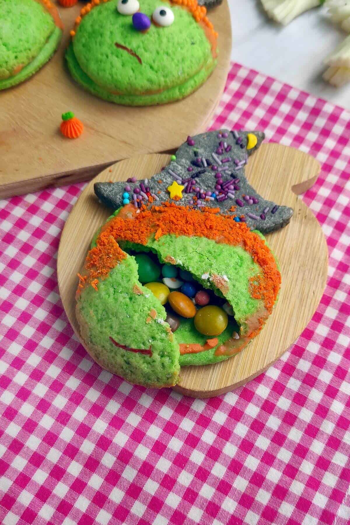A green, monster-themed cookie with orange hair, candy eyes, and a mouth, partially broken open to show colorful candy inside, on a wooden board with a checkered tablecloth underneath.