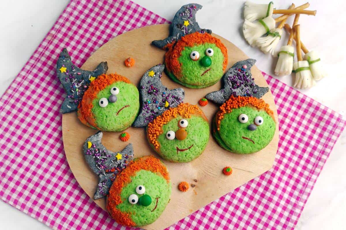 Five green cookies decorated as witches with candy hats, orange sprinkle hair, and candy eyes are arranged on a round wooden board. Edible mushroom decorations are on the side.