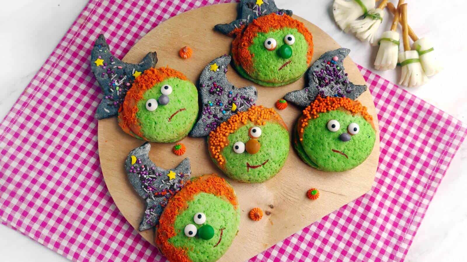 Five green cookies decorated as witches with candy hats, orange sprinkle hair, and candy eyes are arranged on a round wooden board. Edible mushroom decorations are on the side.