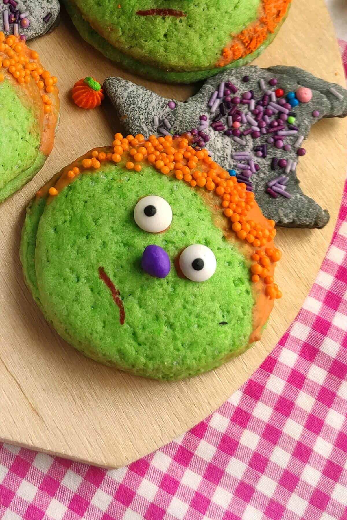 A green cookie decorated as a face with orange sprinkles for hair, candy eyes, and a purple candy nose, next to a bat-shaped cookie with gray icing and sprinkles.