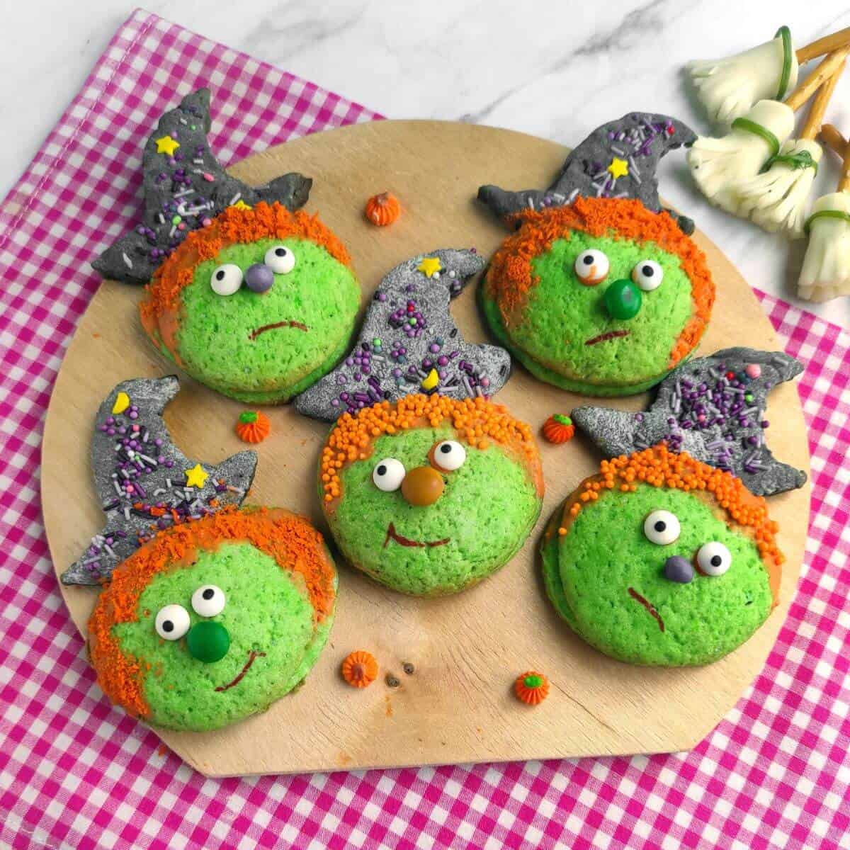 Five green cookies decorated as witch faces with orange candy hair, candy eyes, and pointy hats, arranged on a round wooden board on a pink checkered cloth.