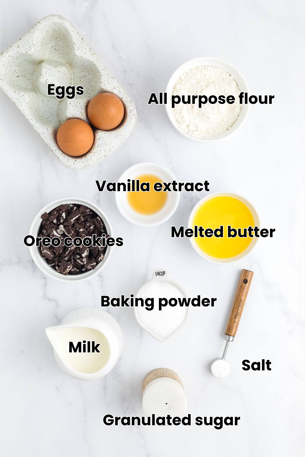 A flat lay of ingredients including eggs, flour, crushed cookies, vanilla, melted butter, milk, sugar, salt, and a measuring spoon on a white surface.