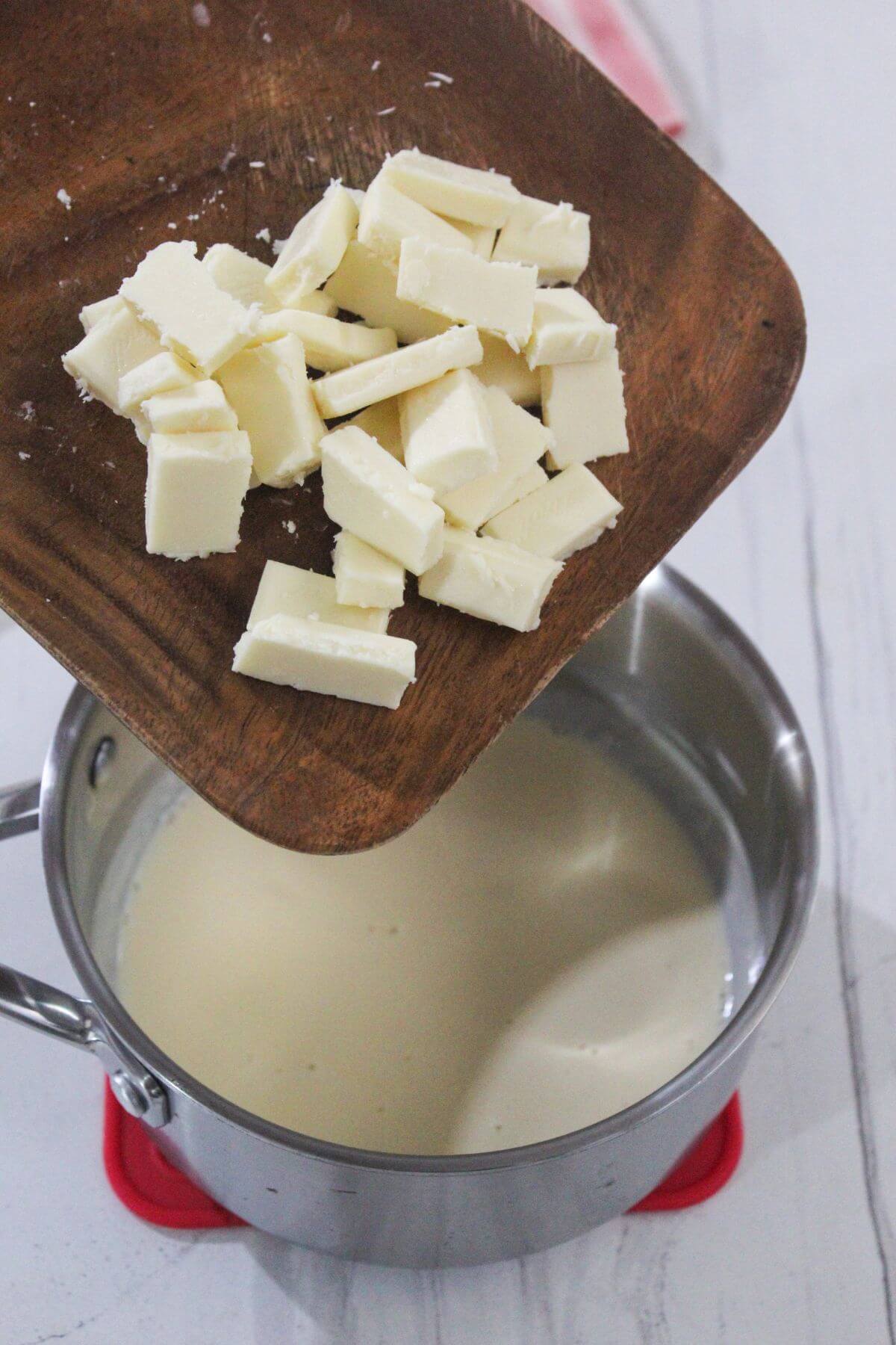 Cubed butter is being added from a wooden board into a saucepan containing a creamy liquid on a white surface.