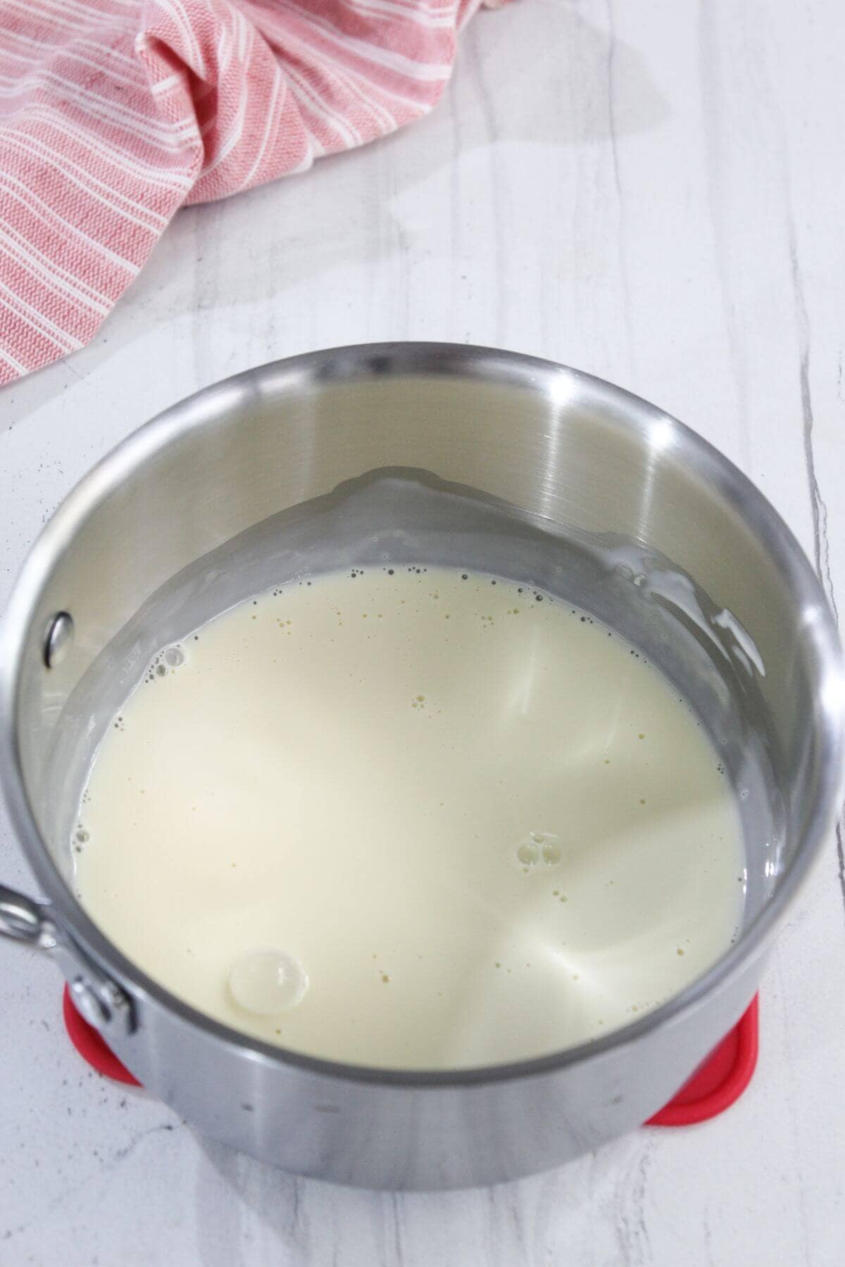 A stainless steel saucepan containing milk sits on a white countertop next to a pink and white striped cloth.