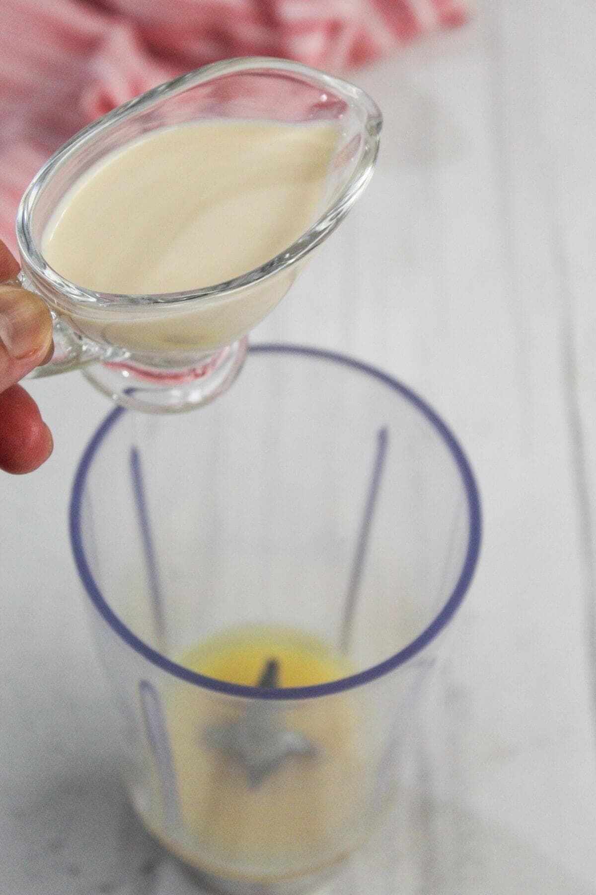 A hand holds a small glass pitcher of cream above a blender cup containing a yellow liquid, ready to pour.