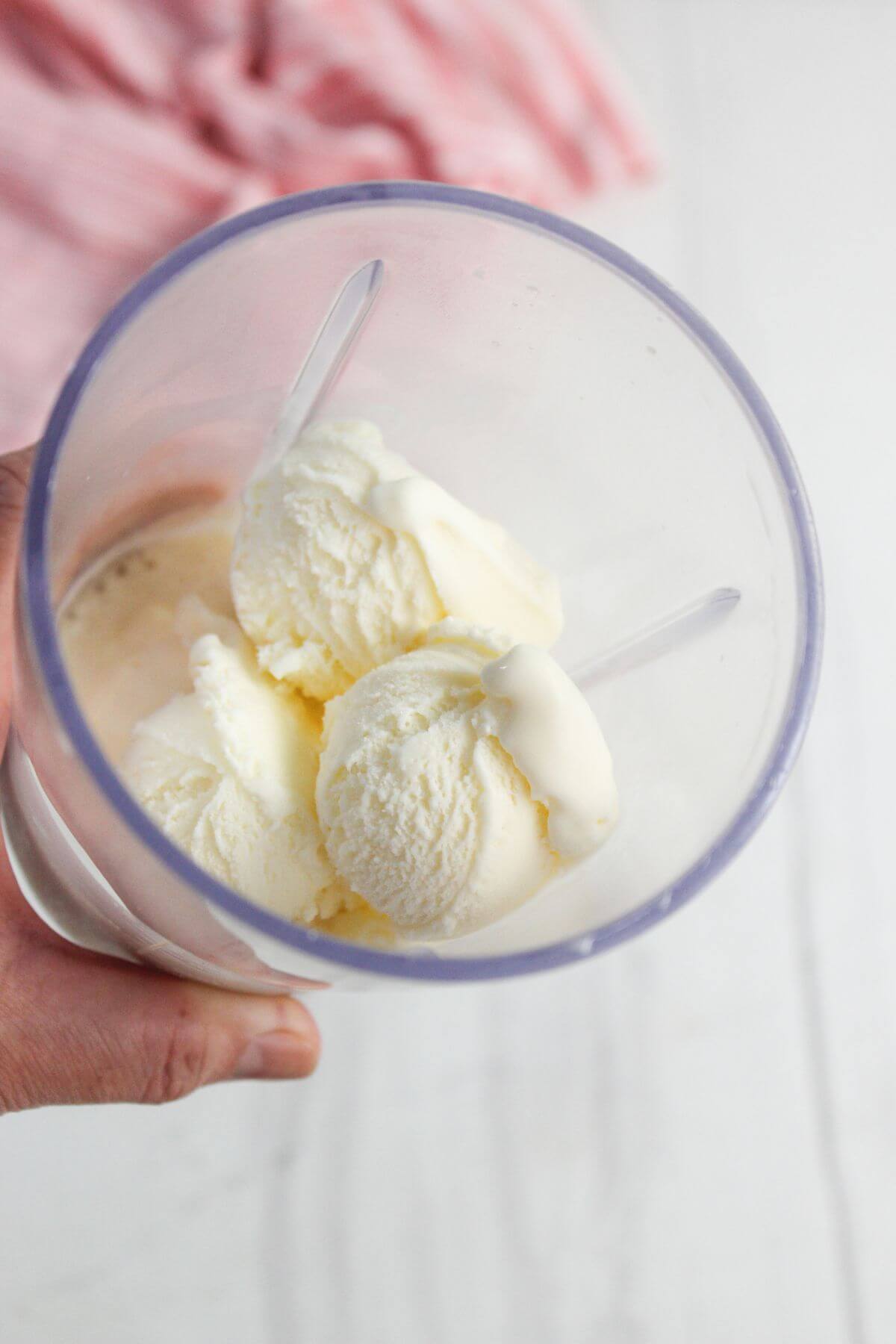 A hand holds a clear glass containing three scoops of vanilla ice cream with two plastic spoons.