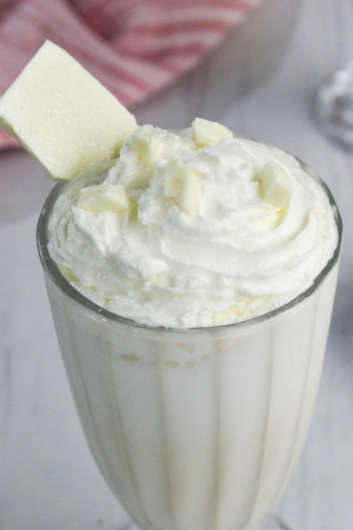 A glass of white chocolate milkshake topped with whipped cream, white chocolate shavings, and a piece of white chocolate.