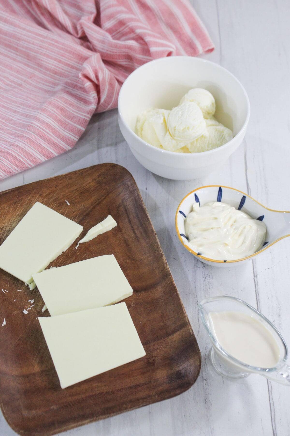 A wooden board with three blocks of cream cheese, a bowl of sliced butter, a small dish of mayonnaise, and a glass jug of cream on a white surface with a pink striped cloth.