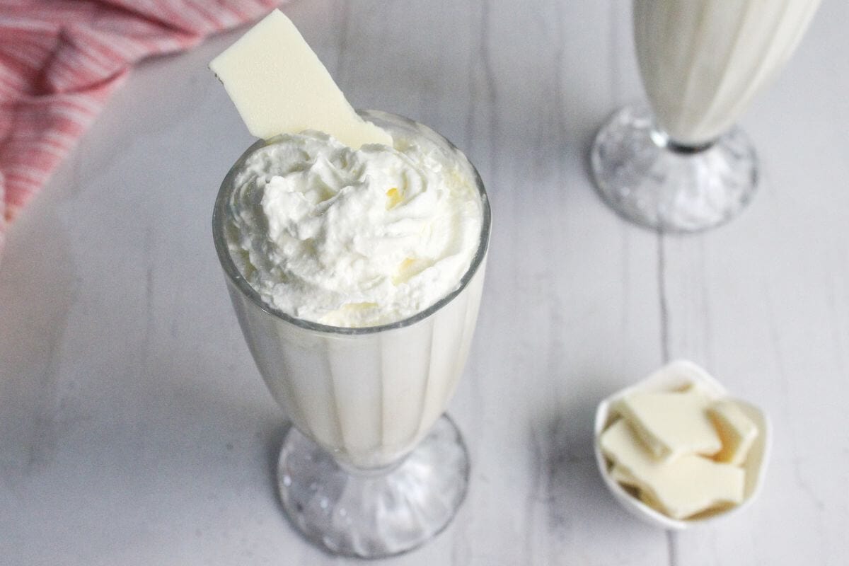 A tall glass of white milkshake topped with whipped cream and a piece of white chocolate, with extra white chocolate pieces in a small dish nearby.
