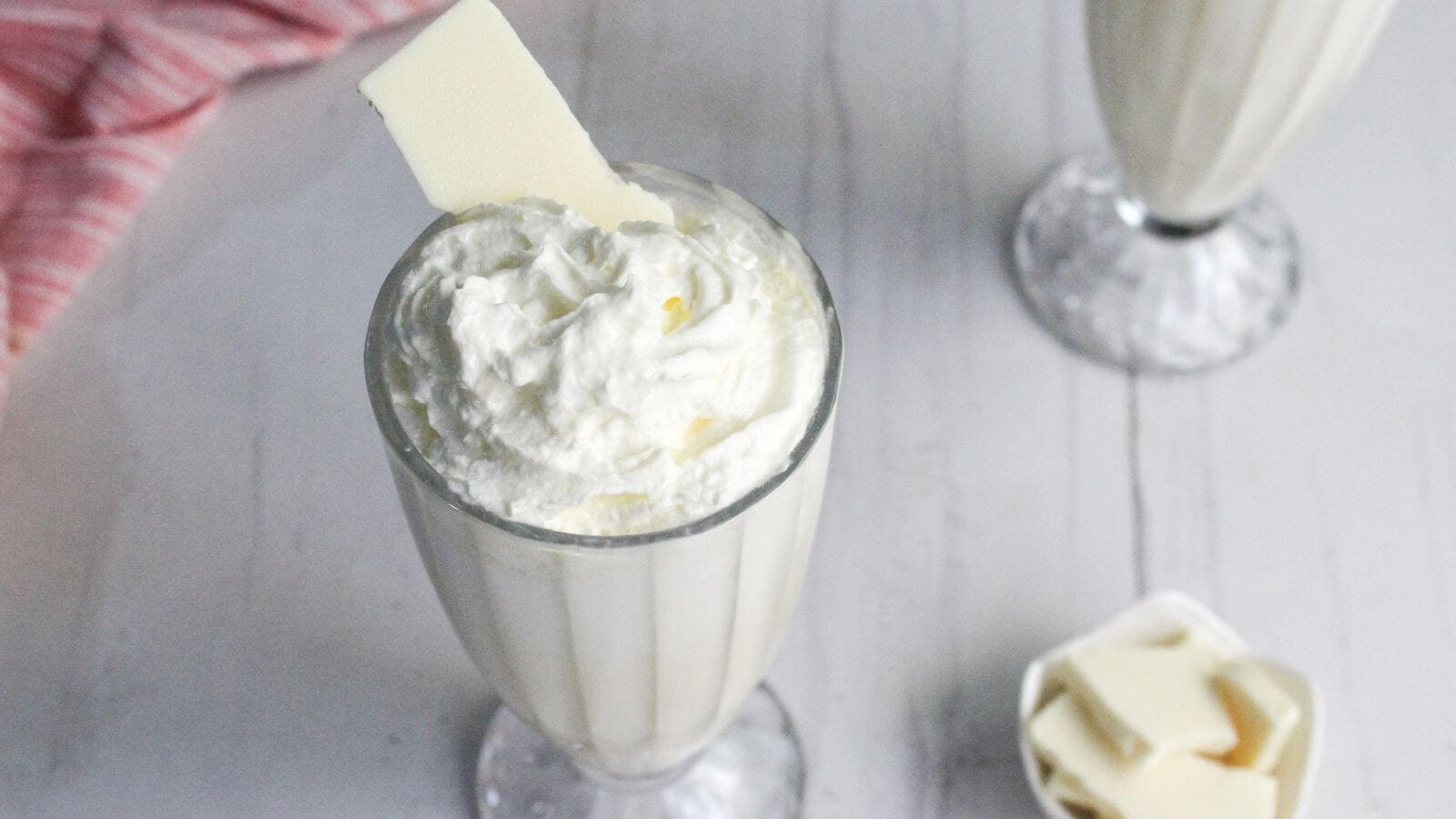 A glass filled with a white milkshake topped with whipped cream and a white chocolate piece, with another milkshake and a bowl of chocolate pieces in the background.