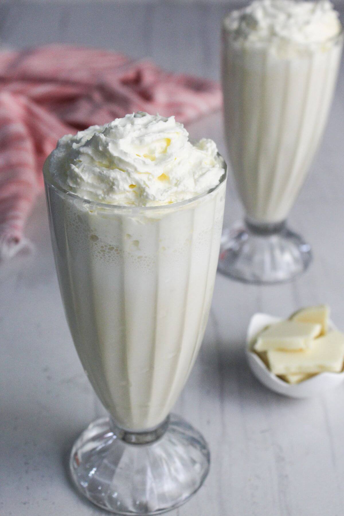 Two tall glasses of white milkshake topped with whipped cream, with a small bowl of white chocolate pieces and a pink cloth in the background.