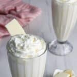Two tall glasses of white chocolate milkshake topped with whipped cream and a white chocolate piece, with a bowl of white chocolate chunks and a striped cloth in the background.