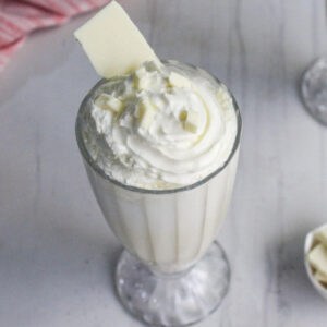 A glass filled with a white milkshake topped with whipped cream, white chocolate shavings, and a piece of white chocolate, on a white surface.