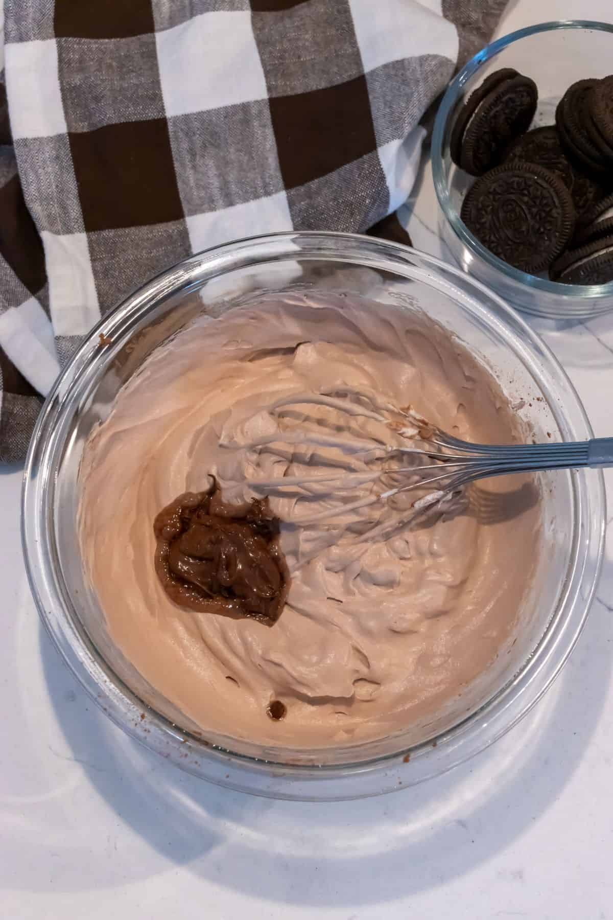 A glass bowl of chocolate Oreo mousse mixture is being whisked, with a spoonful of chocolate spread added. A bowl of sandwich cookies and a checkered cloth appear in the background.