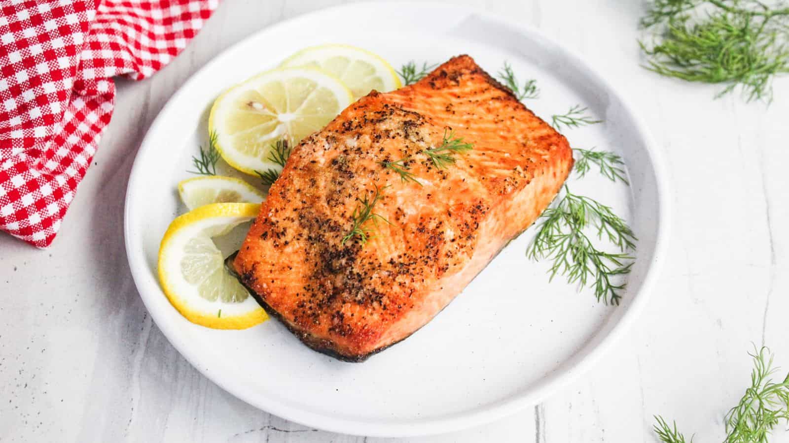 Grilled salmon fillet garnished with dill, served on a white plate with lemon slices, on a white surface with a red checkered cloth.