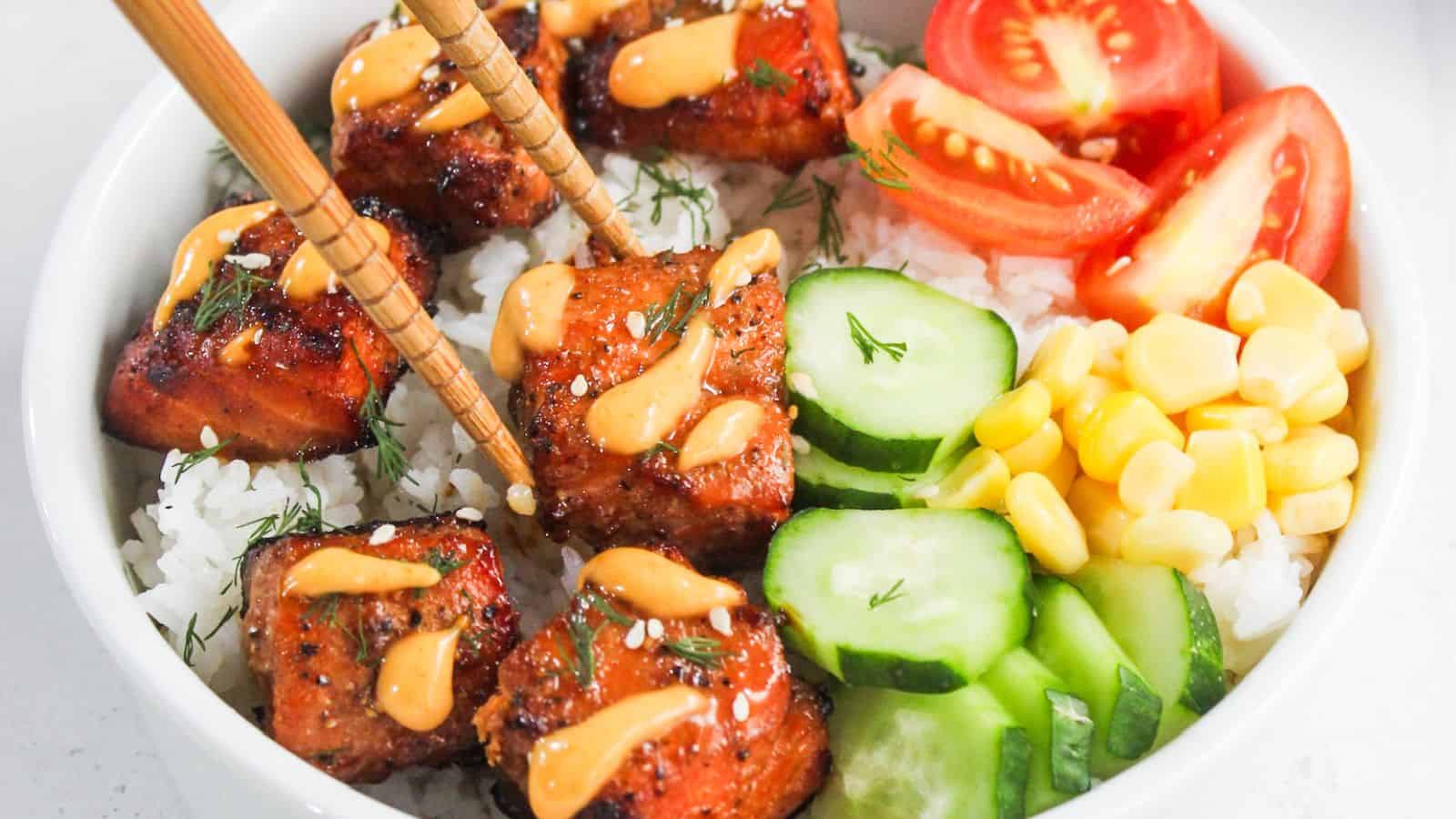 A bowl of white rice topped with glazed salmon cubes, sliced cucumbers, cherry tomatoes, corn, and a drizzle of orange sauce, with chopsticks resting on the salmon.