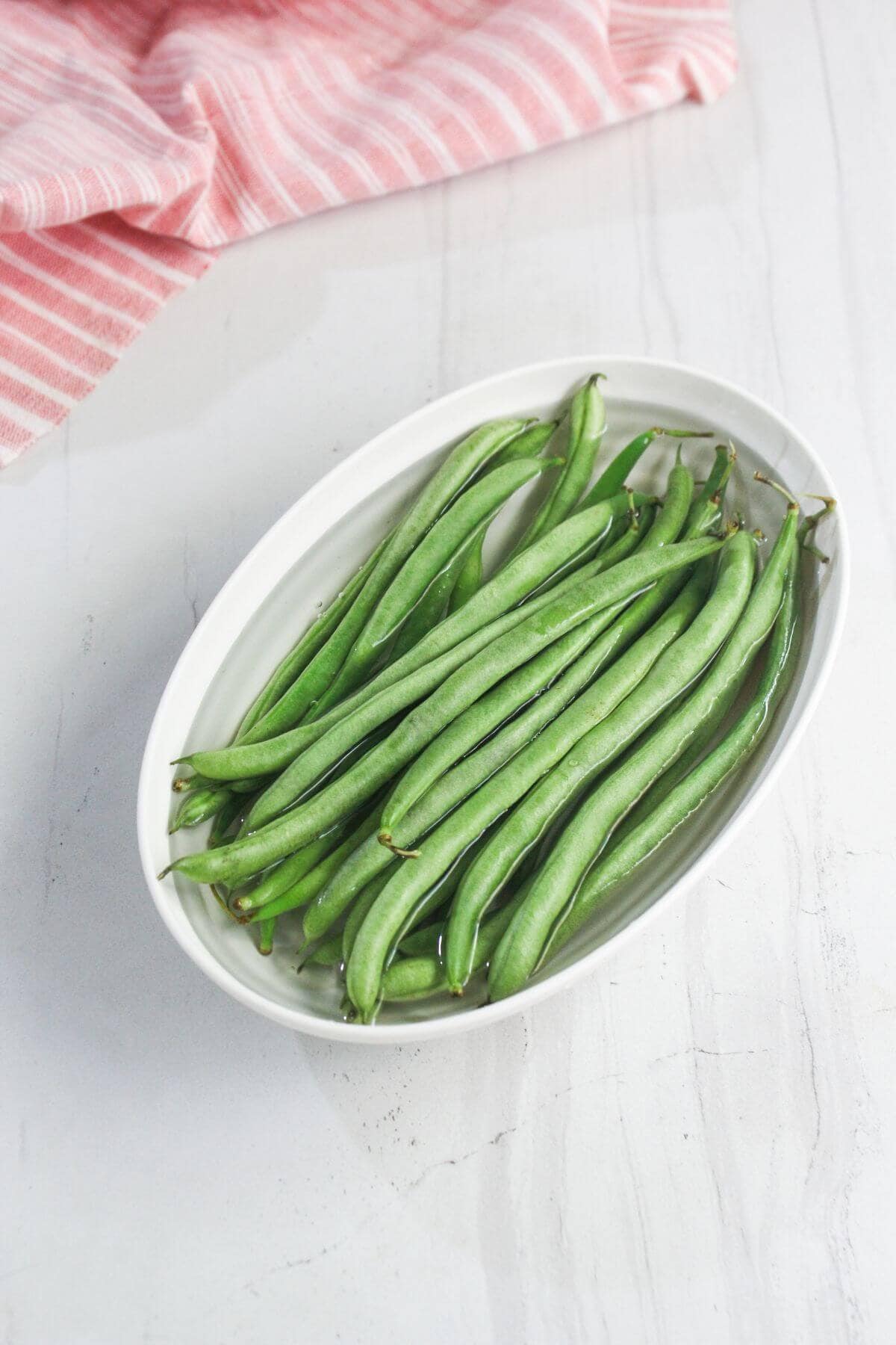 An oval white dish filled with fresh green beans sits on a light marble surface next to a folded pink and white striped cloth.