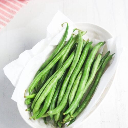A white oval dish lined with paper towels holds fresh green beans, placed on a white surface with a pink striped cloth in the background.