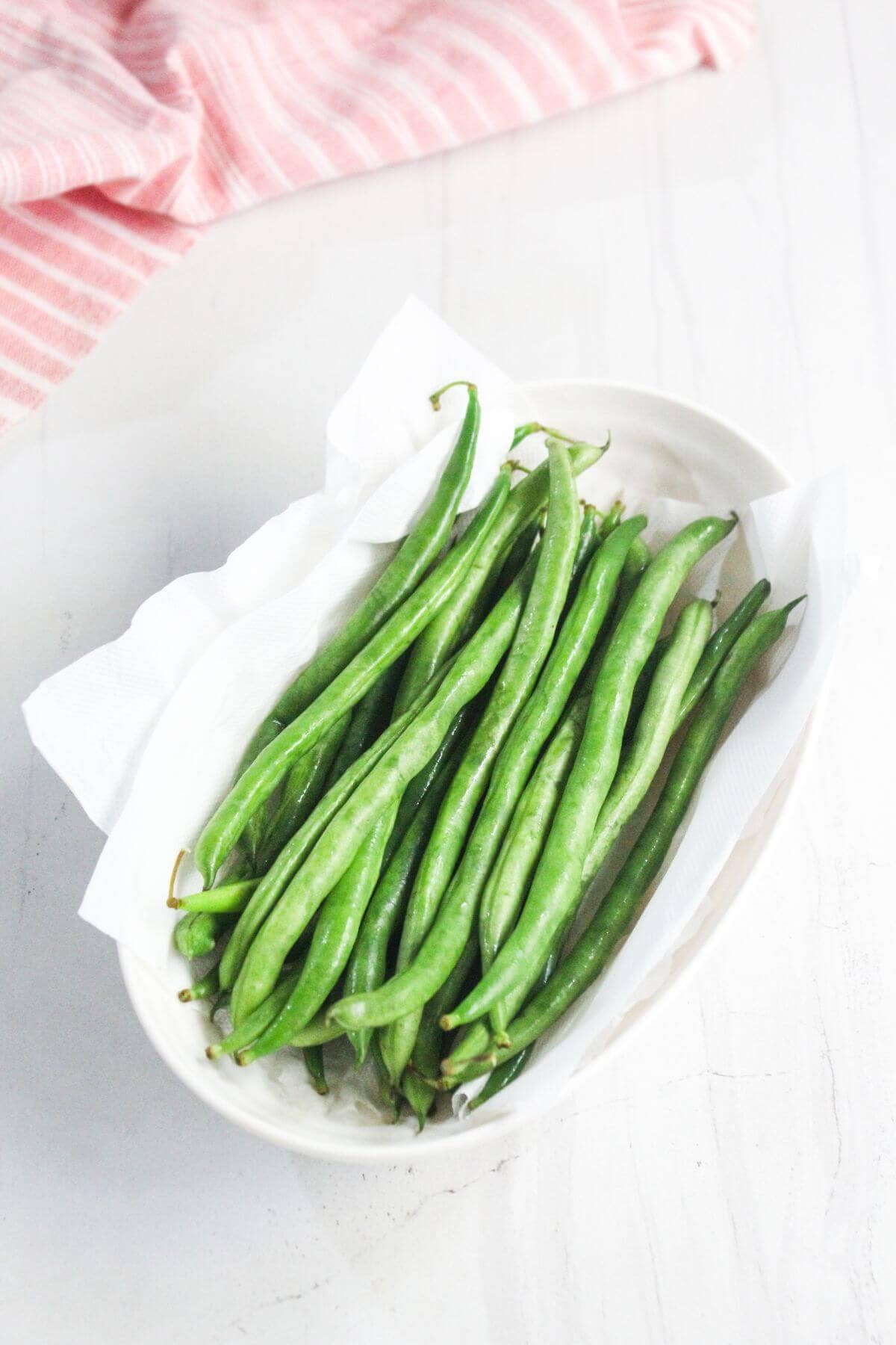 A white oval dish lined with paper towels holds fresh green beans, placed on a white surface with a pink striped cloth in the background.