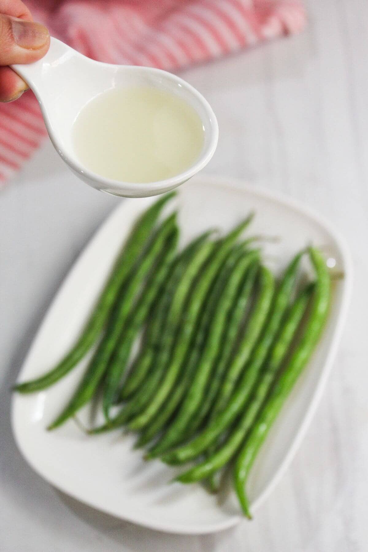A hand holds a small white spoon with liquid above a plate of raw green beans on a white surface.