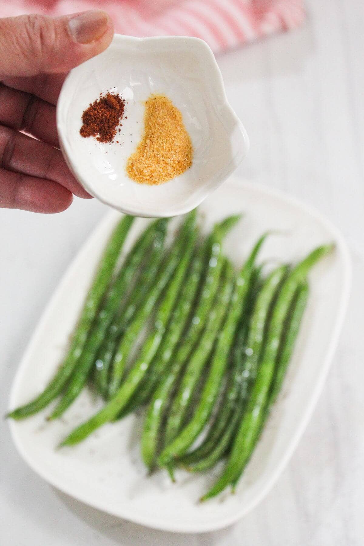 A hand holds a small dish with two piles of spices above a plate of uncooked green beans on a white surface.