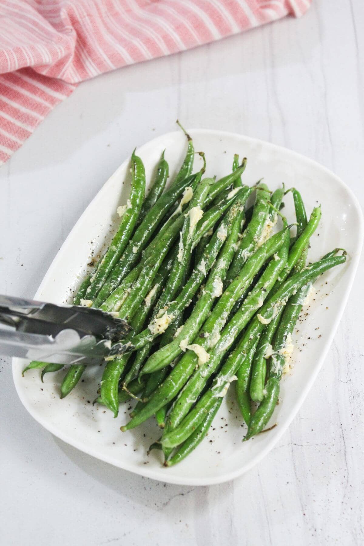 A white plate of cooked green beans topped with grated cheese, with metal tongs on the side, set on a white surface.