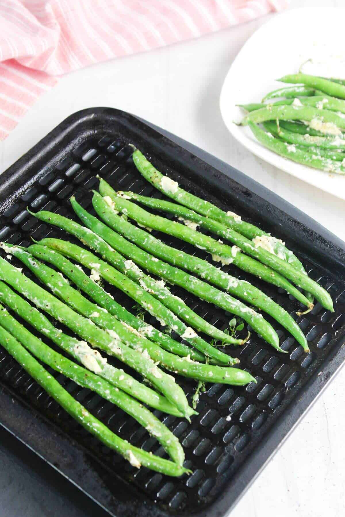 Fresh green beans coated with crumbled cheese and seasoning arranged on a black grill pan, with a plate of green beans in the background.