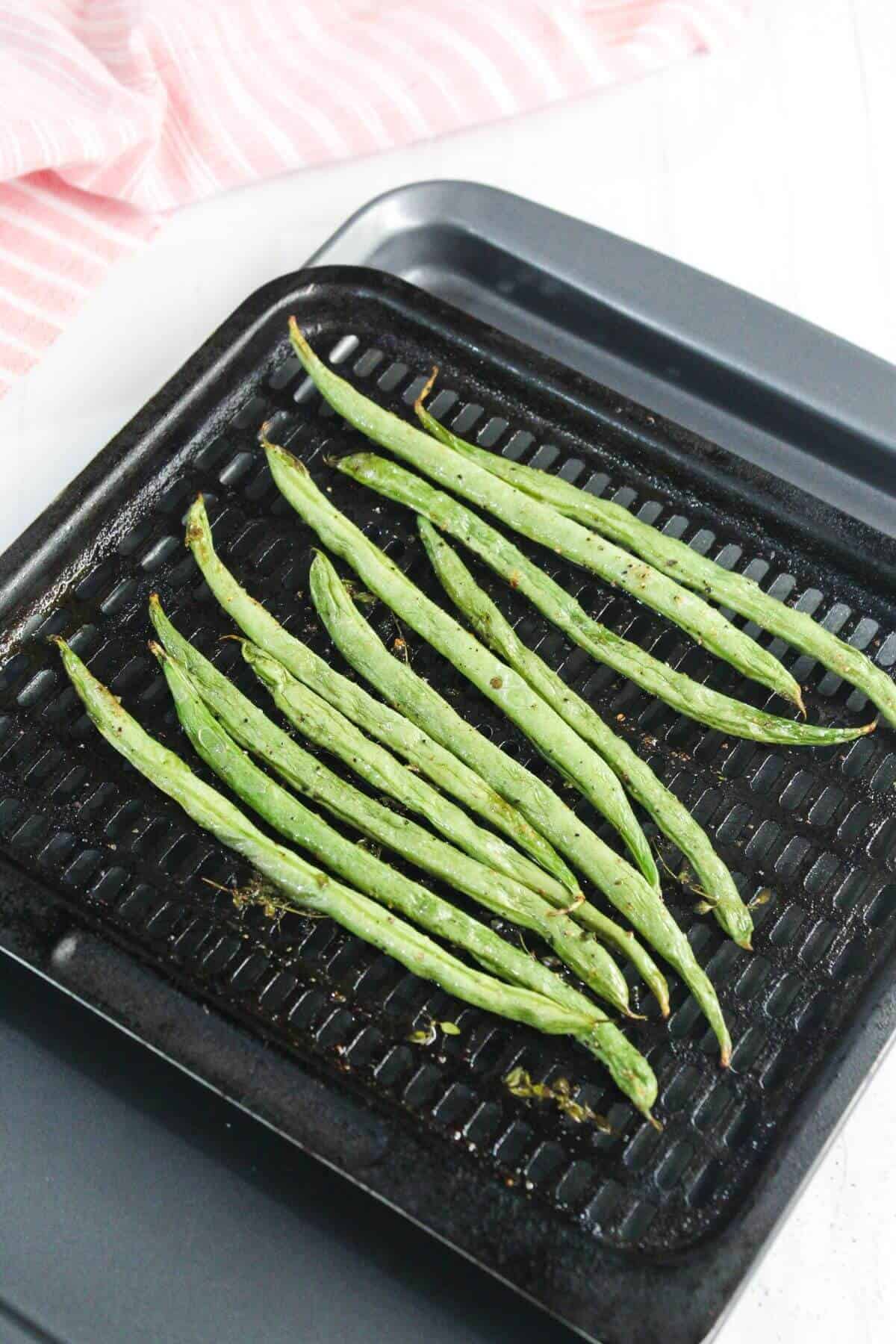 Grilled green beans arranged in a single layer on a black grill tray, with a pink striped cloth in the background.