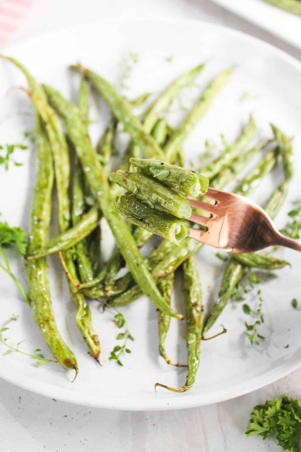 A plate of roasted green beans garnished with herbs, with a fork lifting several beans in the foreground.