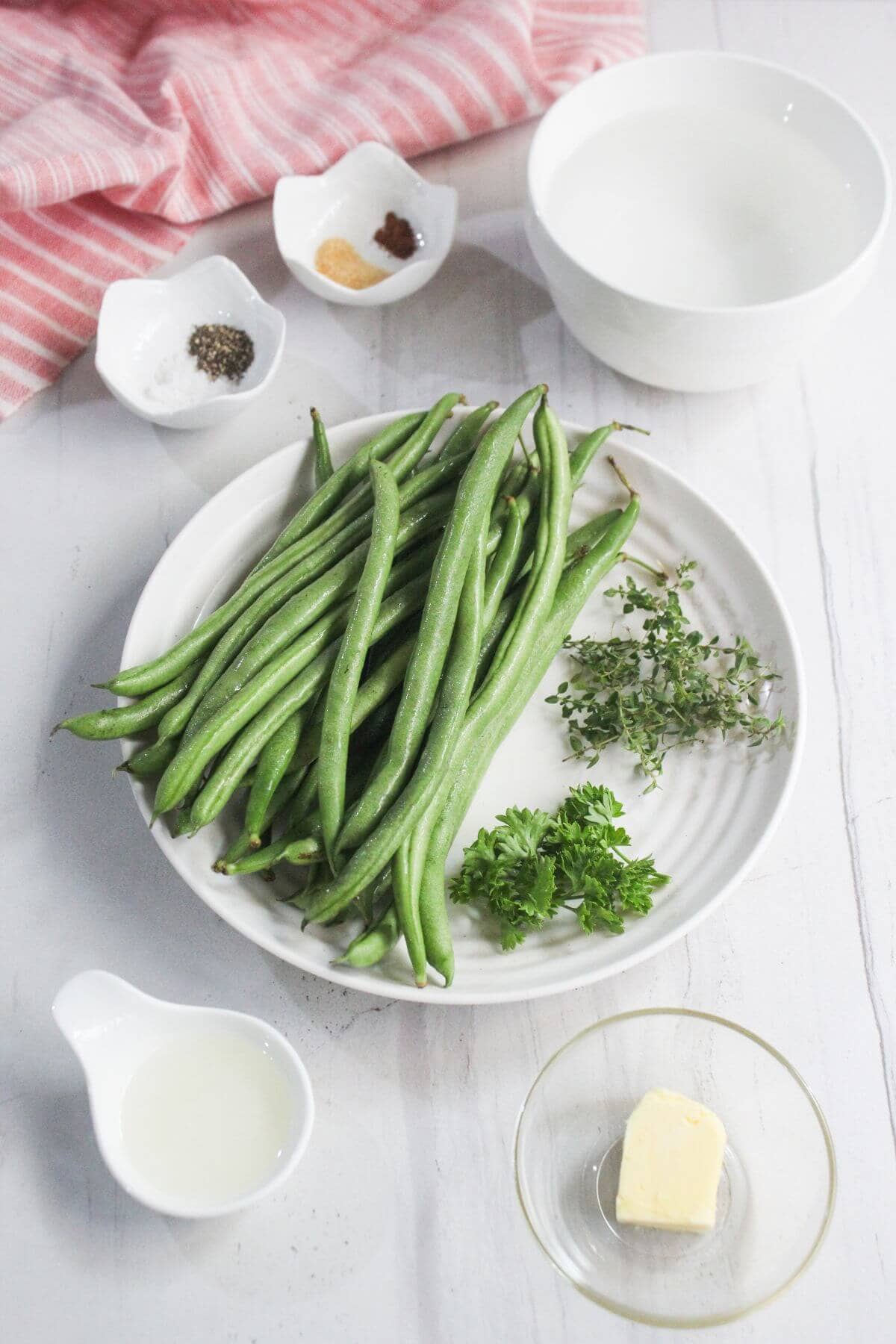 A white plate with fresh green beans, thyme, and parsley, surrounded by small bowls containing butter, oil, spices, and water, on a white surface.