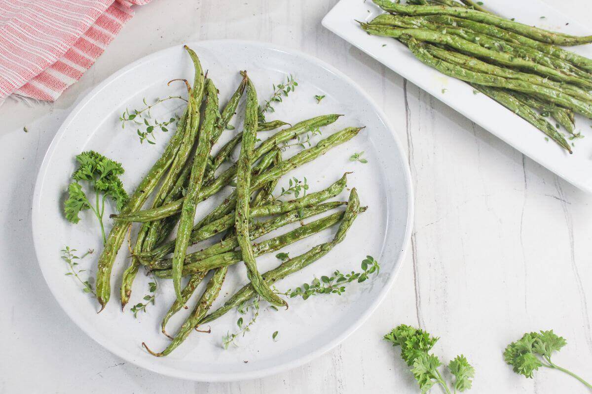 Roasted green beans arranged on a white plate, garnished with fresh herbs, with additional green beans and parsley on the table.