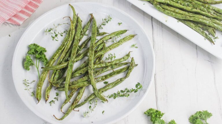 A plate of roasted green beans garnished with fresh parsley and thyme, set on a white table.