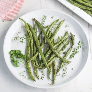 A white plate with roasted green beans garnished with fresh herbs, placed on a light surface.