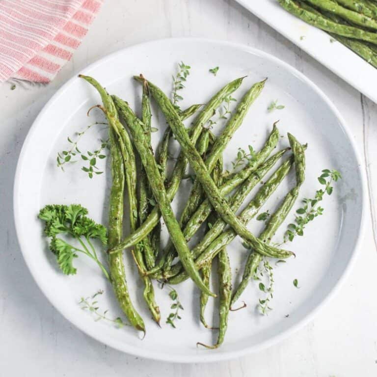 A white plate with roasted green beans garnished with fresh herbs, placed on a light surface.