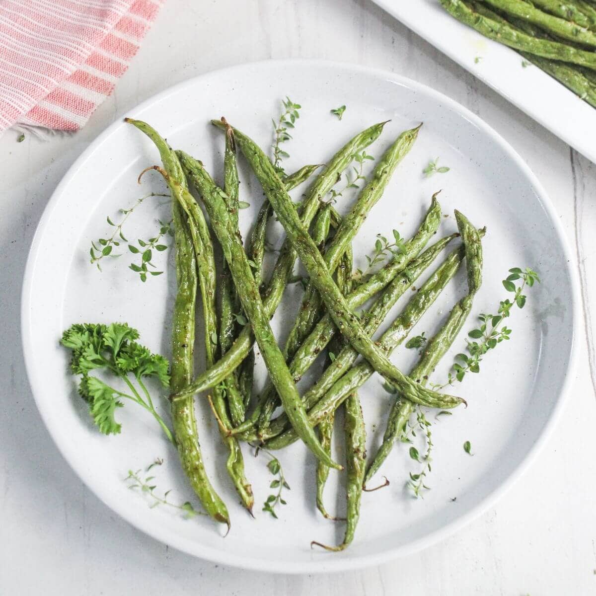 A white plate with roasted green beans garnished with fresh herbs, placed on a light surface.