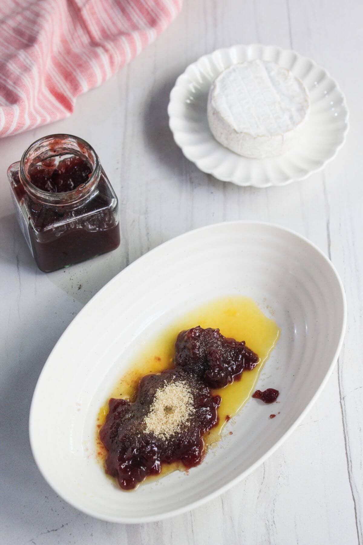 A dish with jam, melted butter, and brown sugar, a jar of jam, and a round of cheese on a plate are arranged on a white countertop.