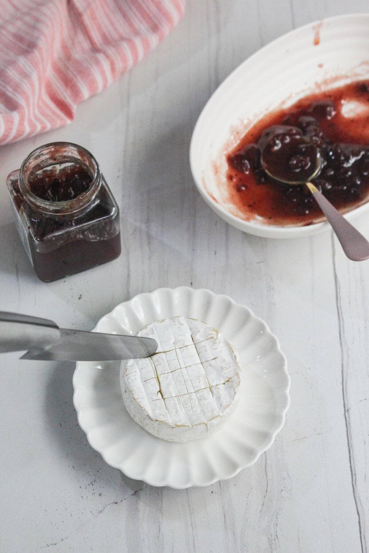 A wheel of brie cheese scored with a knife sits on a white plate, near a jar and bowl of red fruit jam with spoons on a marble surface.