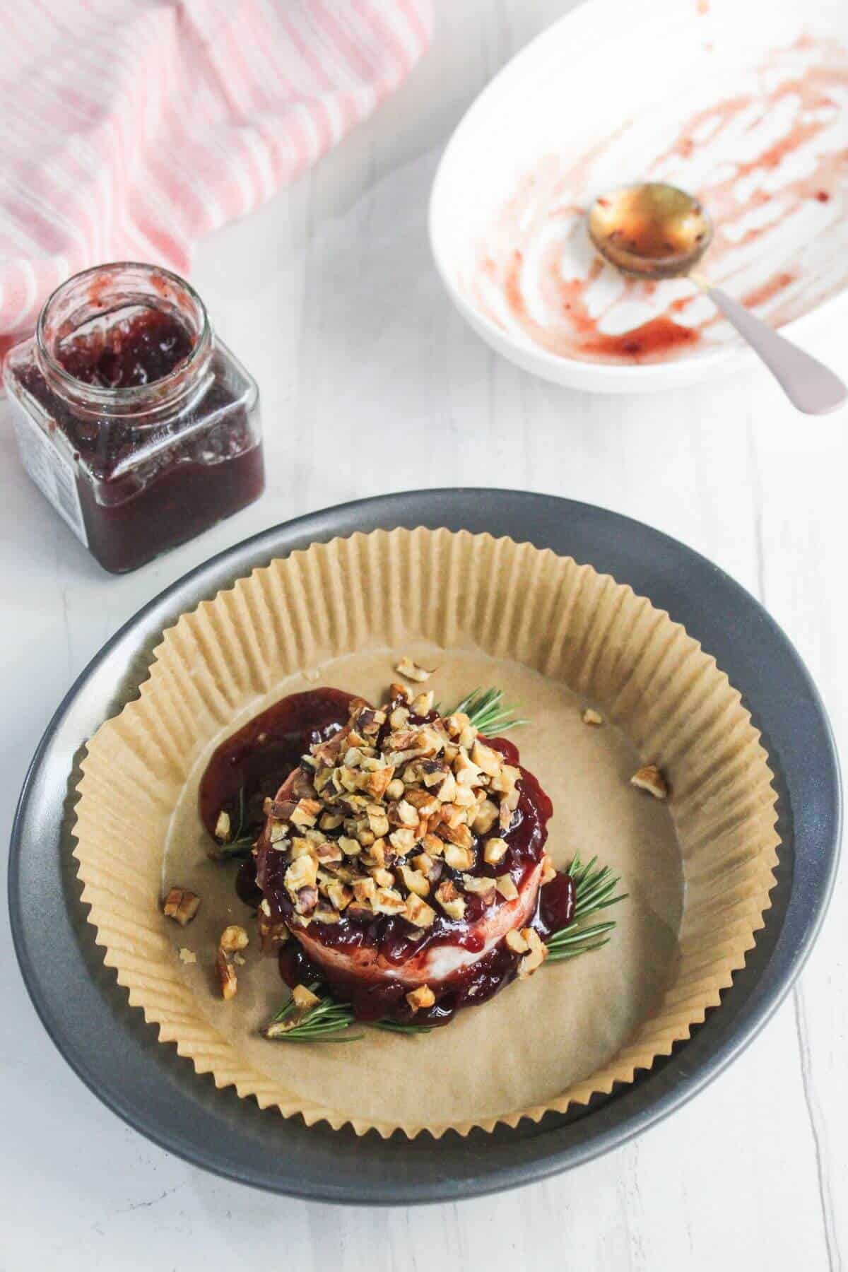 A round baked cheese topped with chopped nuts and fruit preserves sits on parchment paper in a black dish, with a jar of jam and an empty plate nearby.