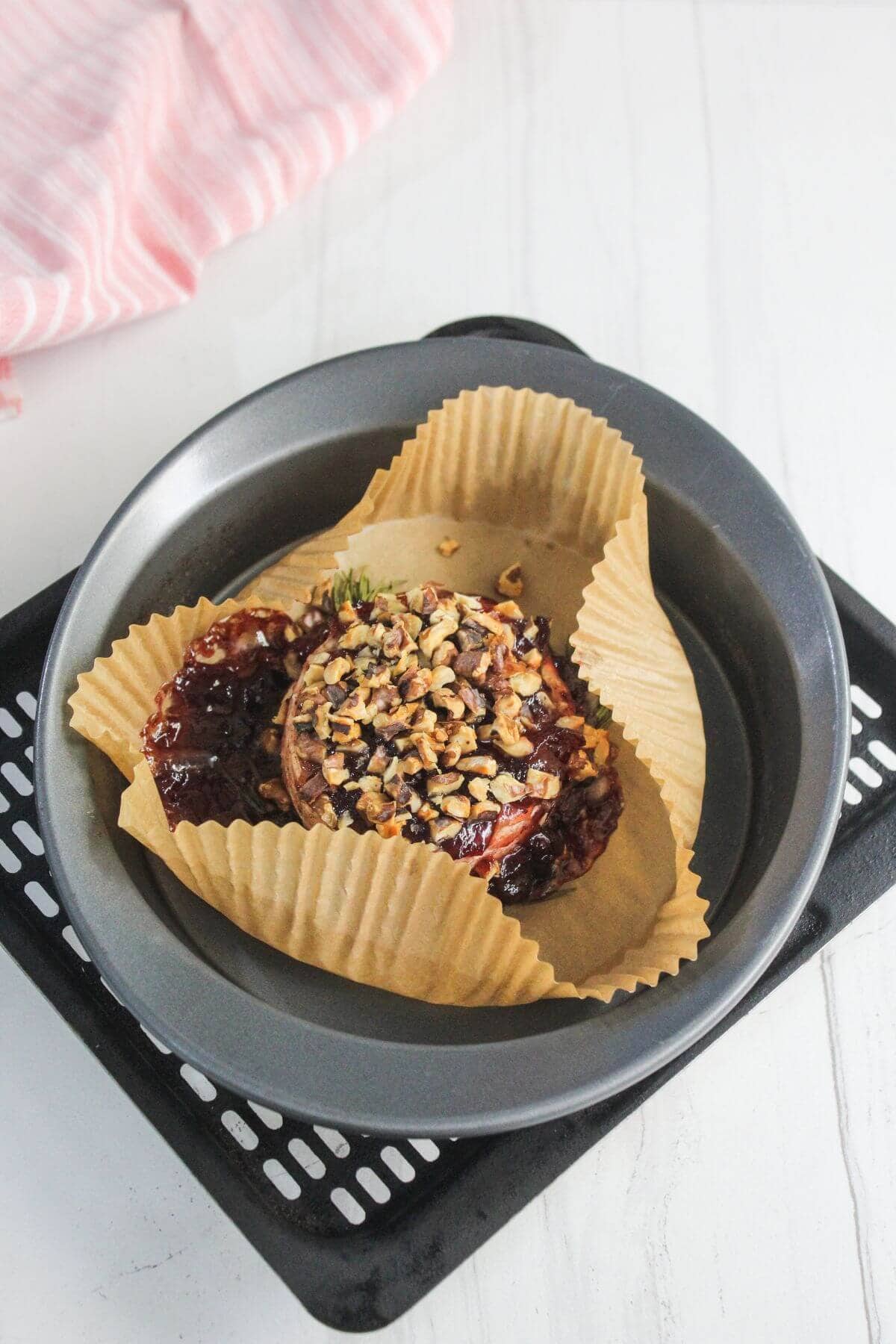 A baked stuffed apple topped with chopped nuts and berry sauce, placed on parchment paper in a round metal baking dish.