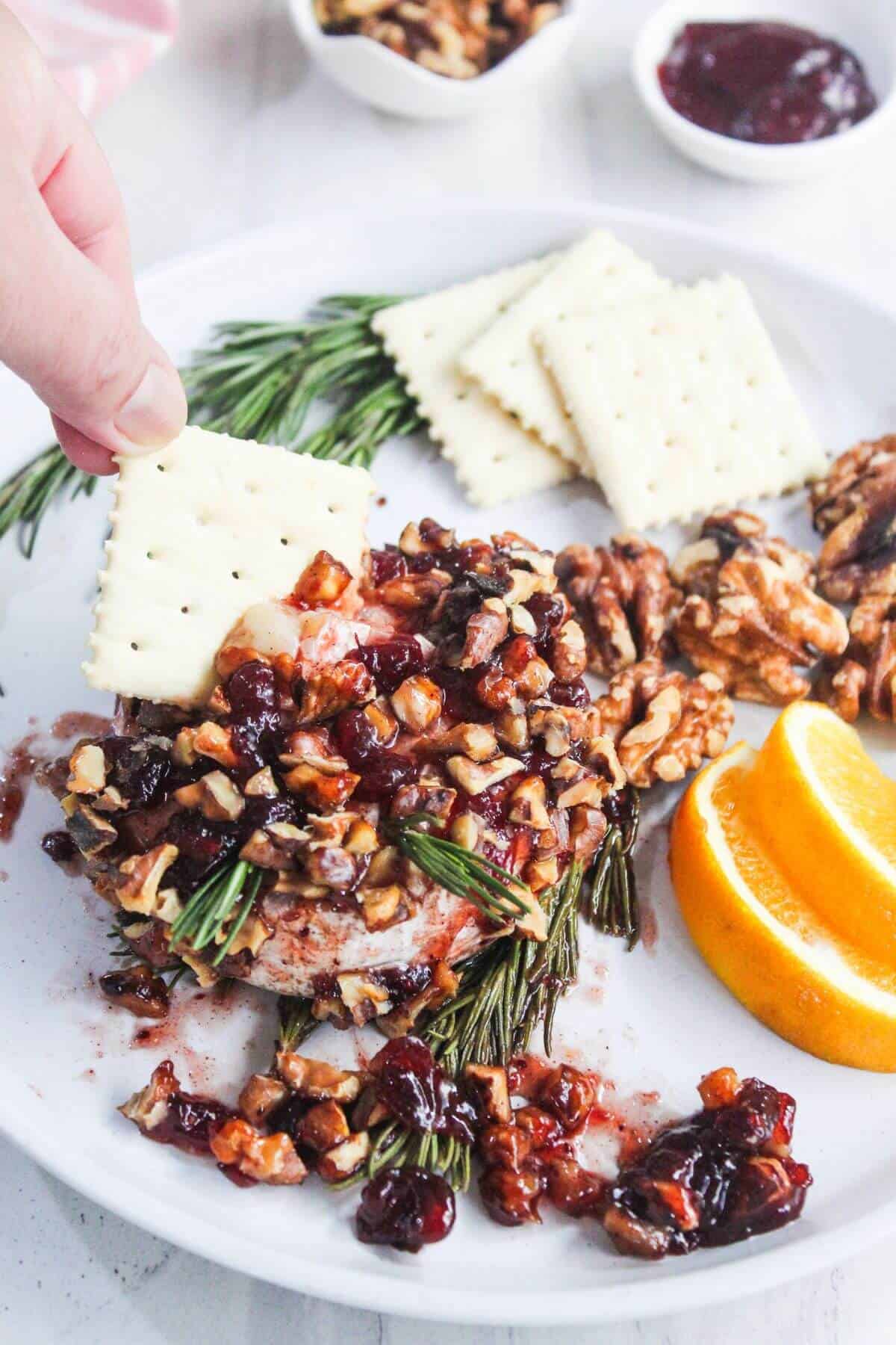 A hand dips a cracker into a cheese wheel topped with chopped nuts and dried fruit, garnished with rosemary, orange slices, whole walnuts, and more crackers on a white plate.