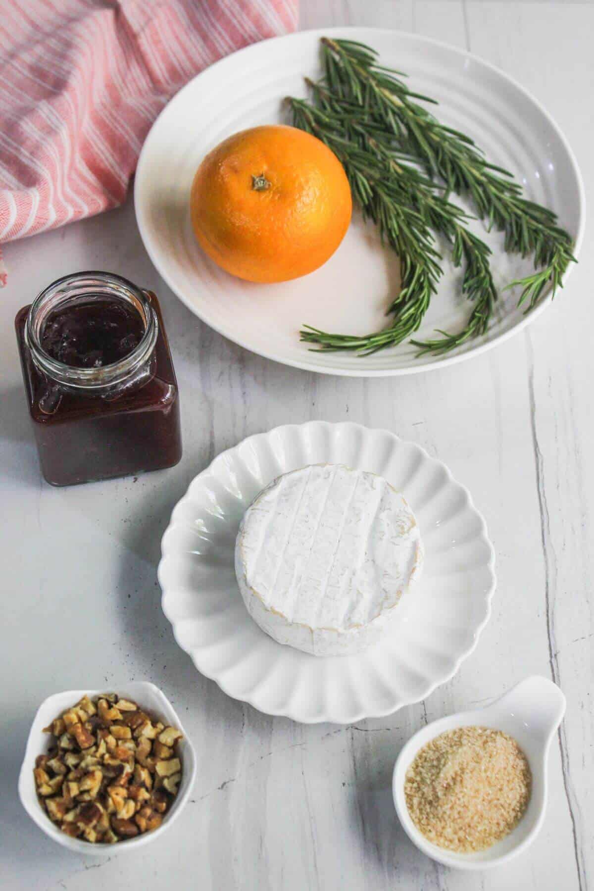 A plate with fresh rosemary and an orange, a jar of jam, a wheel of cheese, chopped nuts, and breadcrumbs arranged on a white surface.