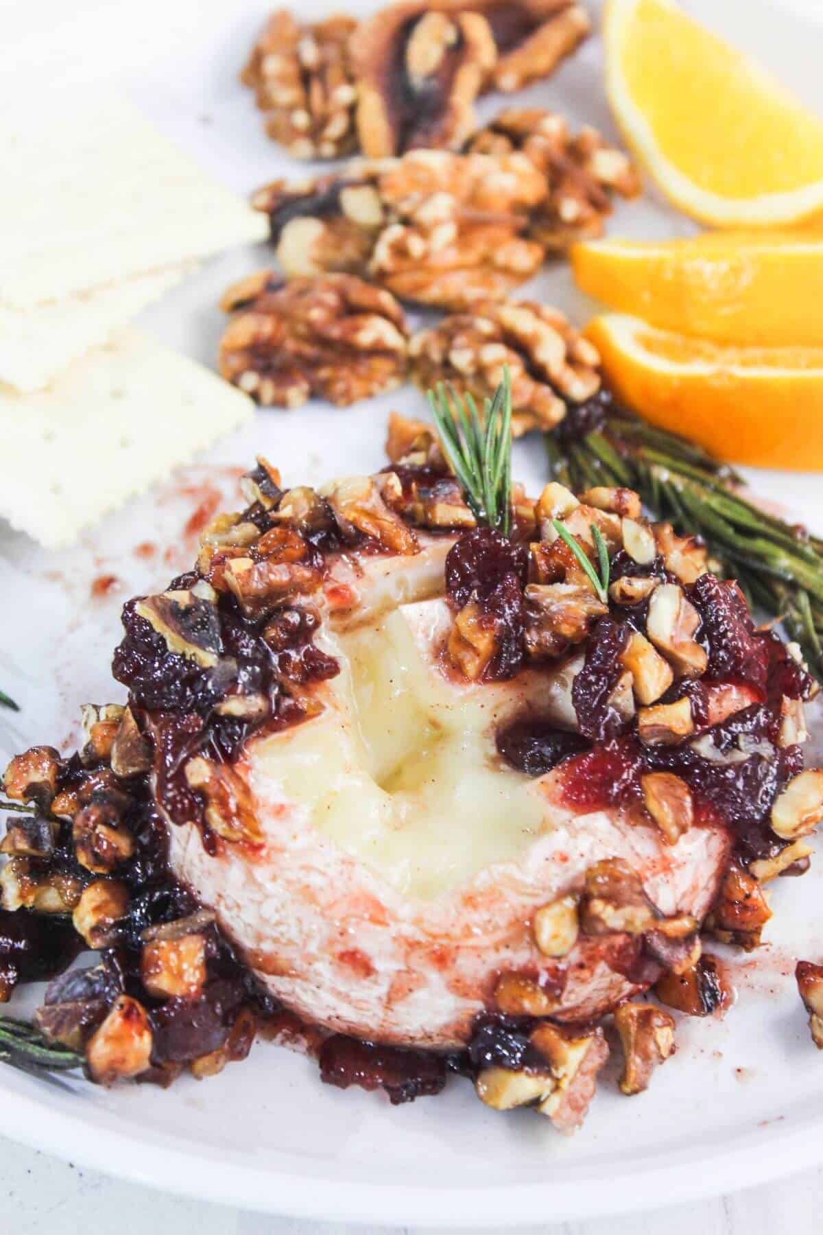 Baked brie topped with chopped nuts and dried fruit, garnished with rosemary, served with orange slices, walnuts, and crackers on a white plate.