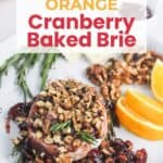 A baked brie topped with chopped nuts and cranberry sauce, garnished with rosemary and orange slices, on a white plate.