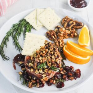 A white plate with crackers, walnuts, orange slices, sprigs of rosemary, and baked brie topped with chopped nuts and dried fruit. A small bowl of jam is in the background.